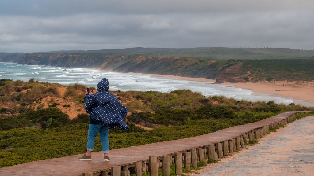 Person im Regenponcho macht Bild mit Smartphone an der Atlantikküse bei Bordeira