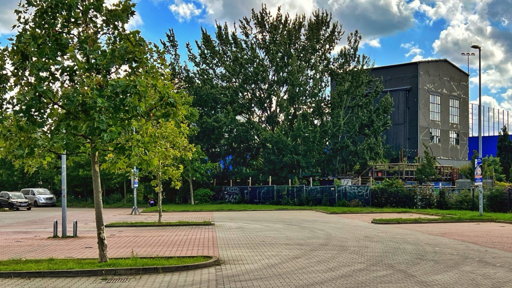 Parkplatz mit Bäumen, im Hintergrund ein kleines Gebäude