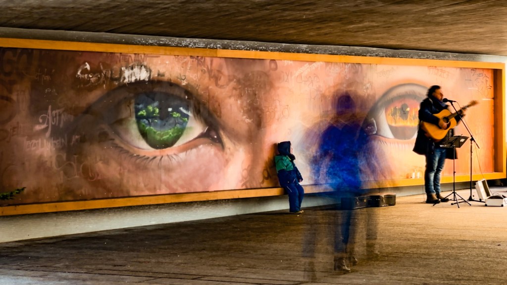 Straßenmusiker in einer Unterführung. Dahinter ein Wandbild mit 2 großen Augen