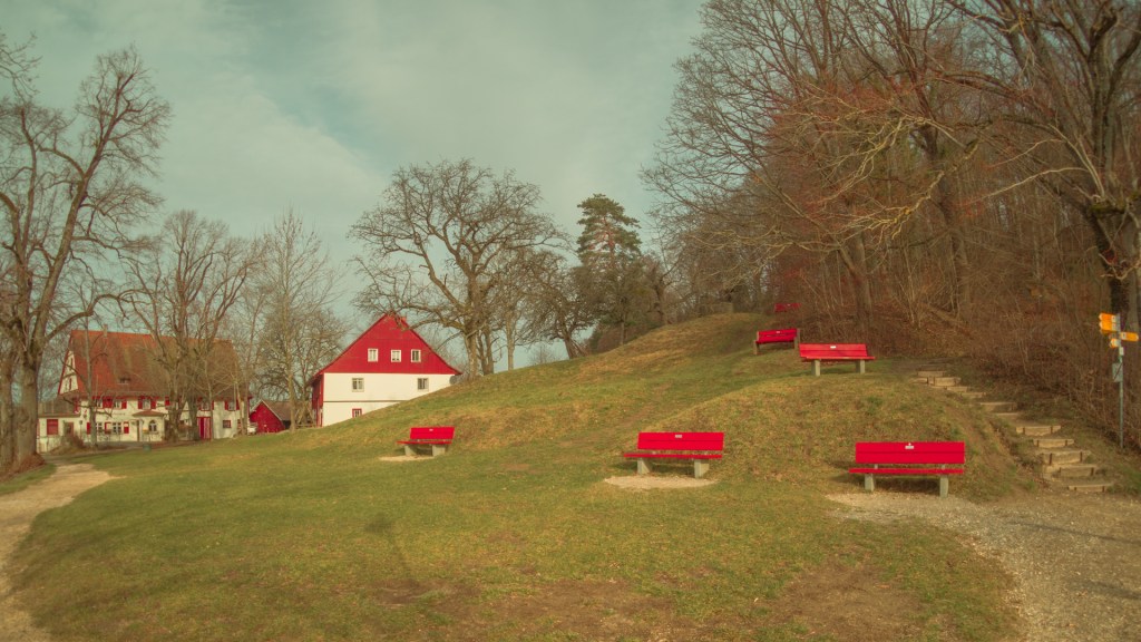 Rote Bänke an einem Hügel, im Hintergrund zwei Häuser