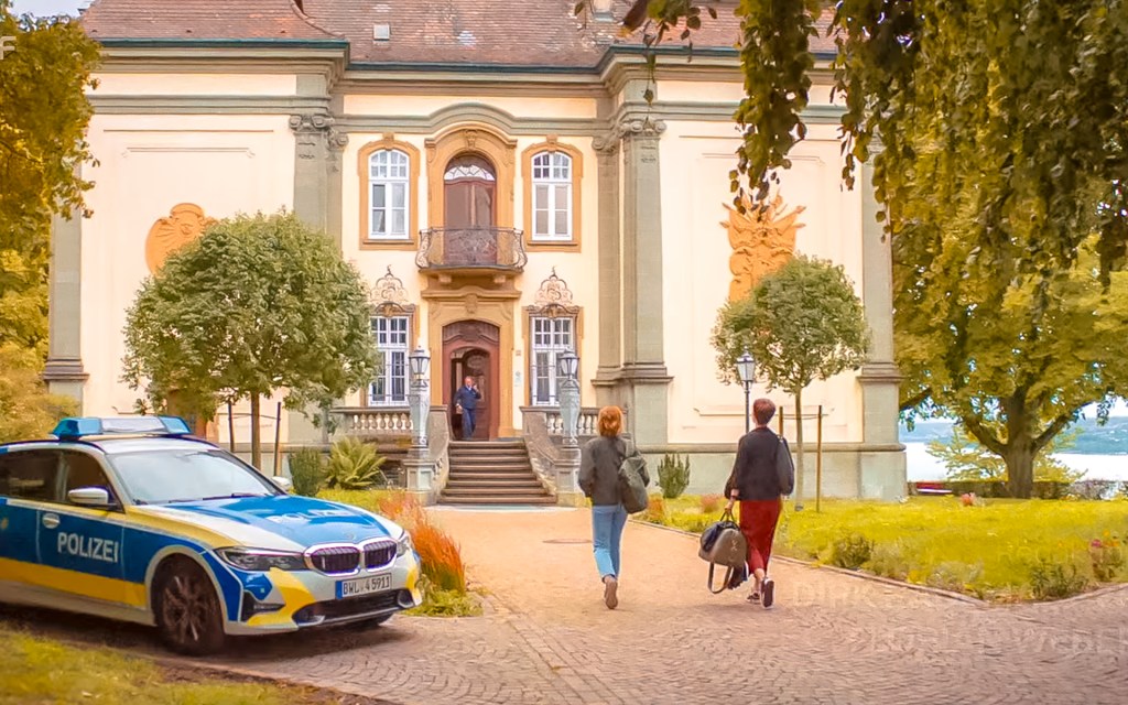 Zwei Personen laufen auf ein Gebäude zu, links steht ein Polizeiauto.