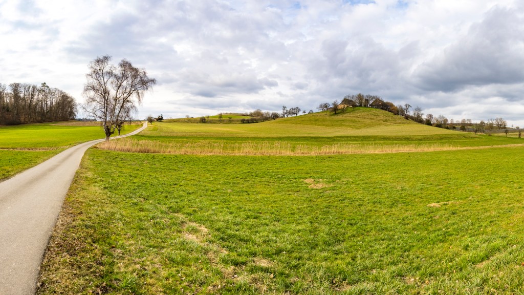 Blick auf eine Landschaft mit einem Hügel auf dem ein Gehöft zu sehen ist. Ein Weg führt von links unten ins Bild.