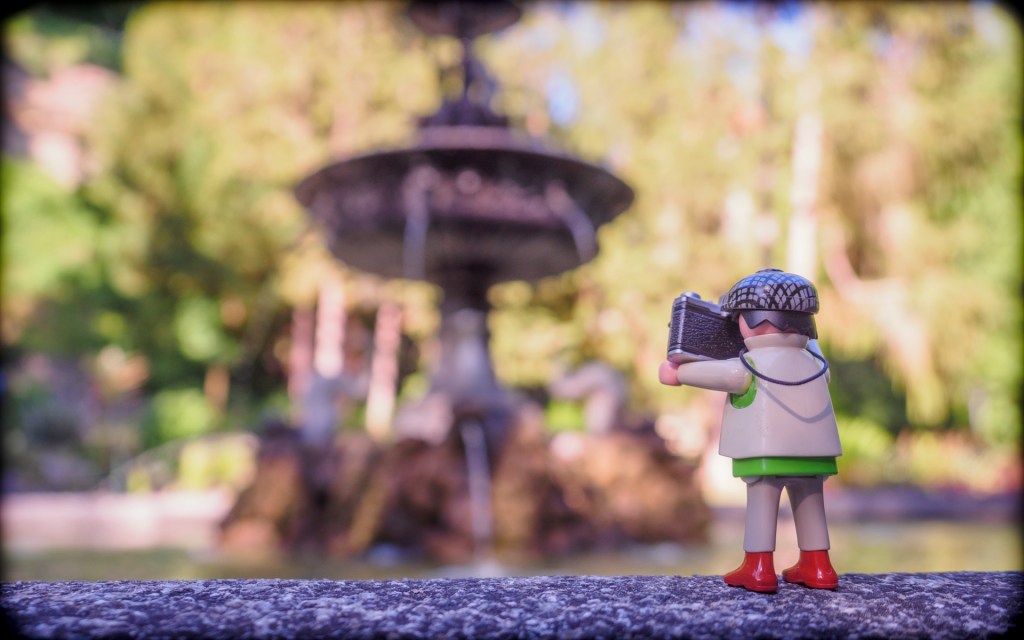 Eine Playmobilfigur steht auf einem Brunnenrand, im Hintergrund in der Unschärfe ein Brunnen