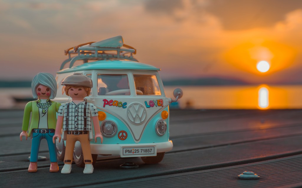 Ein Playmobil VW Bus T1, dafür stehen 2 Figuren. Dahinter geht die Sonne im Bodensee unter.