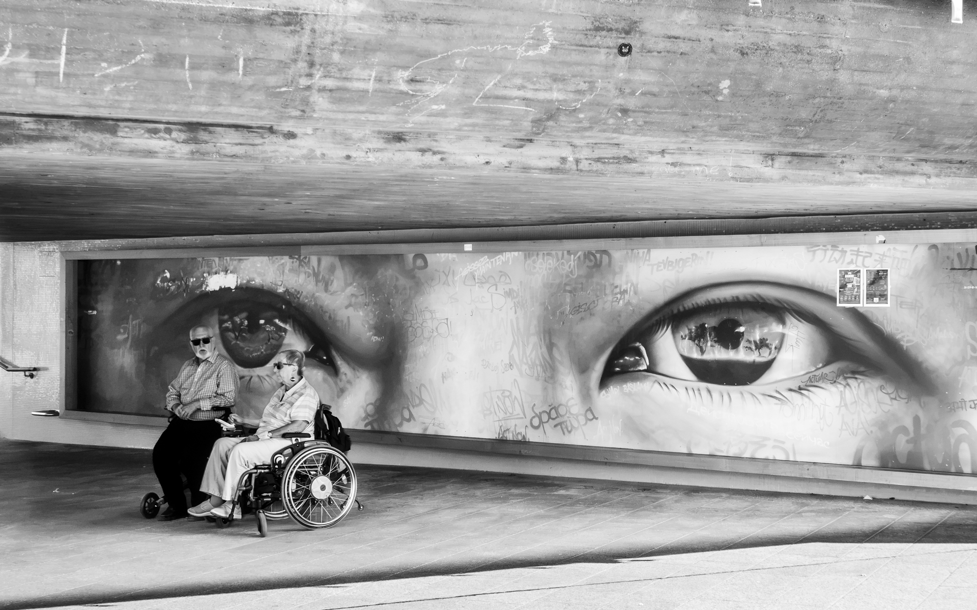 Älterer Mann und Frau im Rollstuhl sitzen unter einer Unterführung. Im Hintergrund an der Wand ein Graffito mit zwei großen Augen.